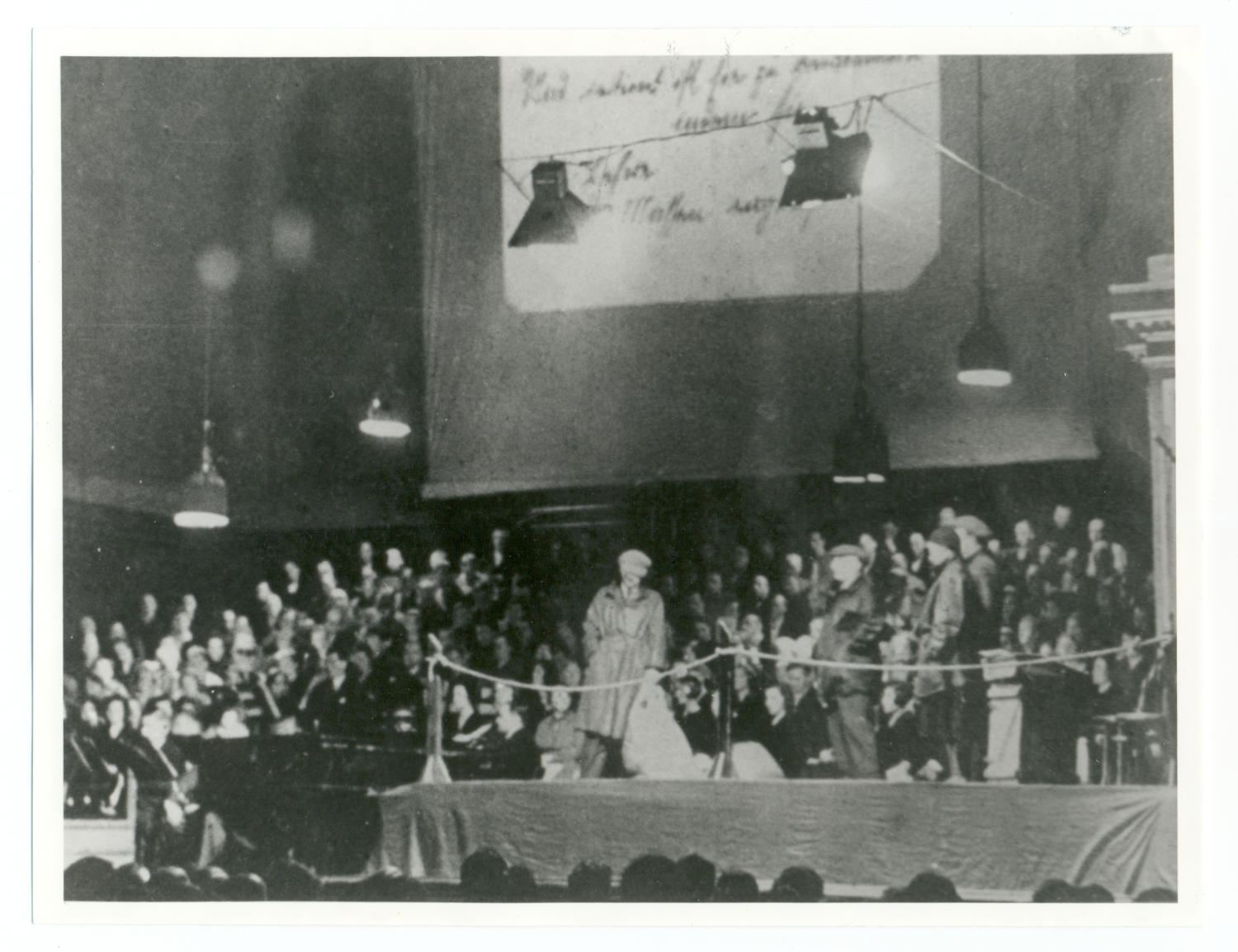 Auf der schwarz-weiß Fotografie sieht man eine abgekordelte Bühne mit vier Männern in Jacken und Mänteln. Viel Publikum sitzt um die Bühne. Im Hintergrund ist ein handgeschriebener Zettel auf eine Leinwand projiziert.