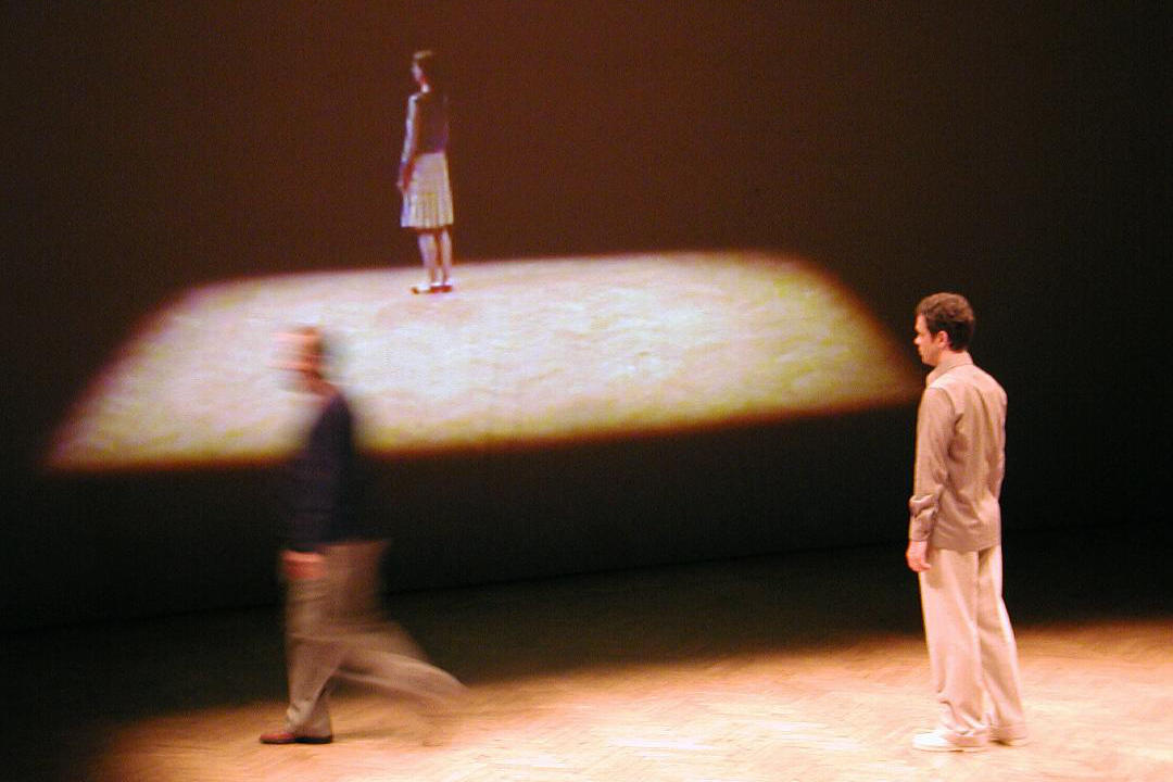 Auf einer Bühne steht rechts ein Mann, schräg mit dem Rücken zur Fotografin. Links ist einer durch rasche Bewegungen nur verschwommen zu sehen. Im Hintergrund befindet sich eine Projektion derselben Bühne mit einer ebenfalls seitlich stehenden Frau.