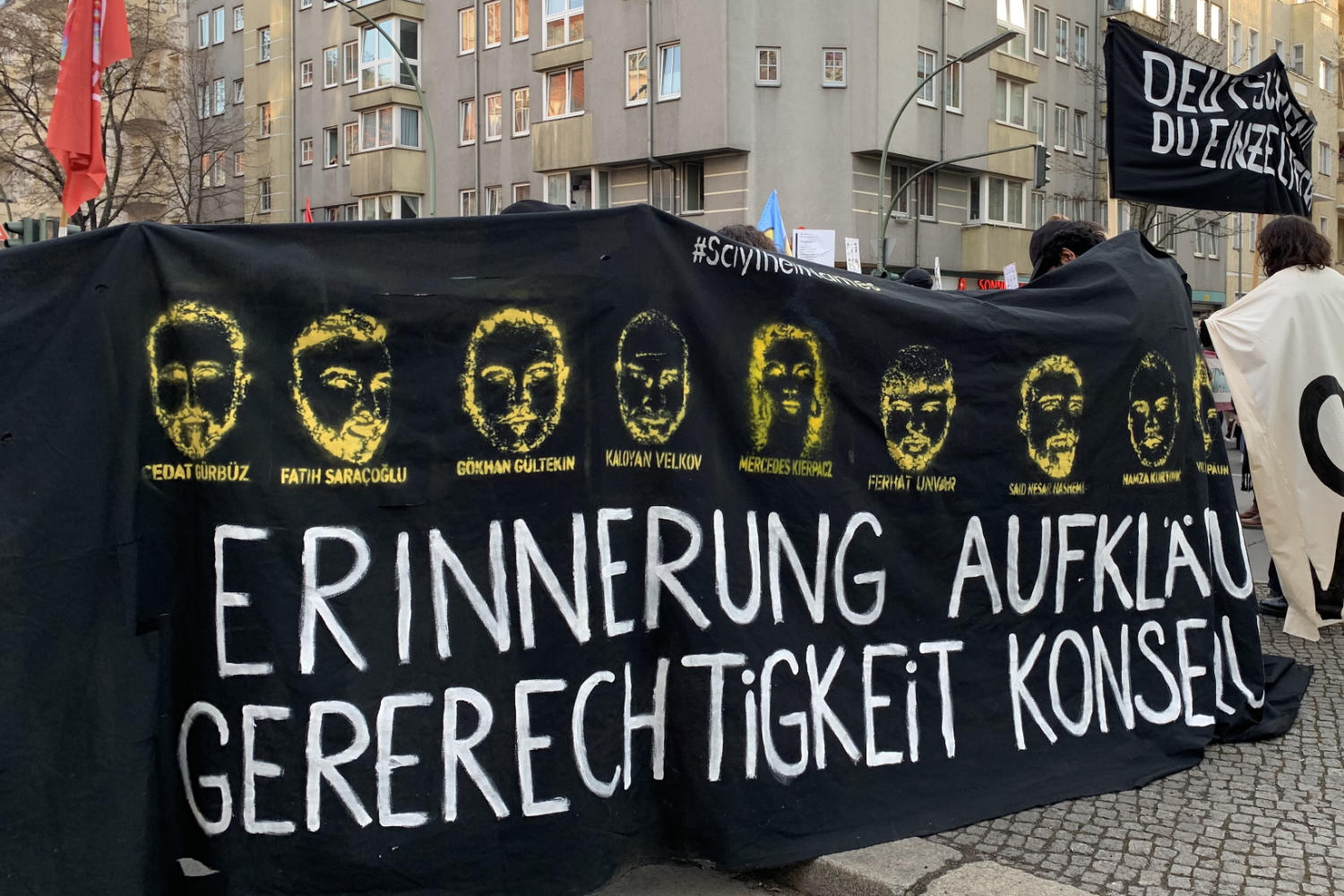 Demonstrierende halten ein schwarzes Banner mit graphischen Darstellungen der Gesichter der Opfer des Anschlags von Hanau am 19.02.2020, ihren Namen und der Aufschrift "Erinnerung, Aufklärung, Gerechtigkeit".