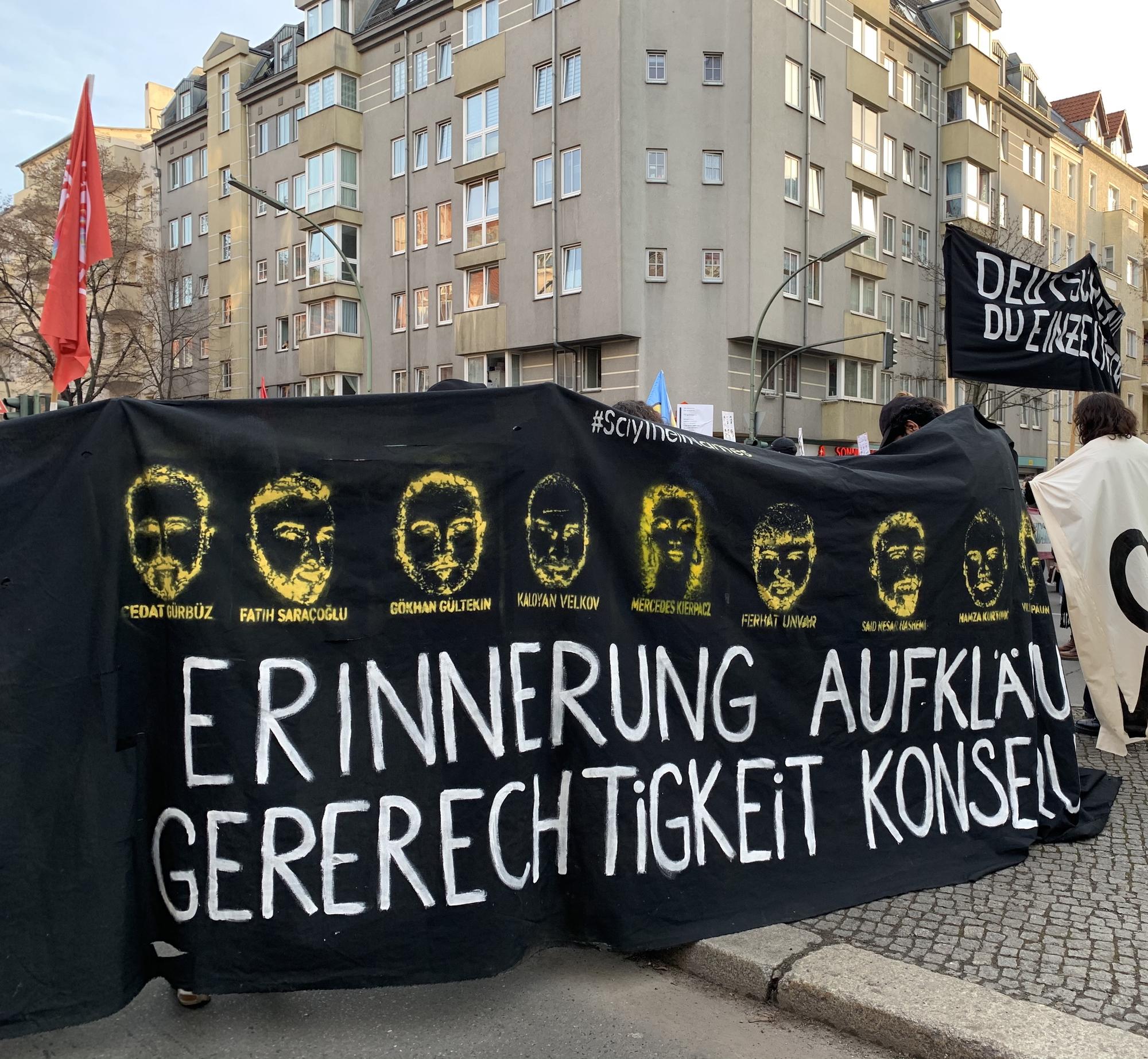 Demonstrierende halten ein schwarzes Banner mit graphischen Darstellungen der Gesichter der Opfer des Anschlags von Hanau am 19.02.2020, ihren Namen und der Aufschrift "Erinnerung, Aufklärung, Gerechtigkeit".