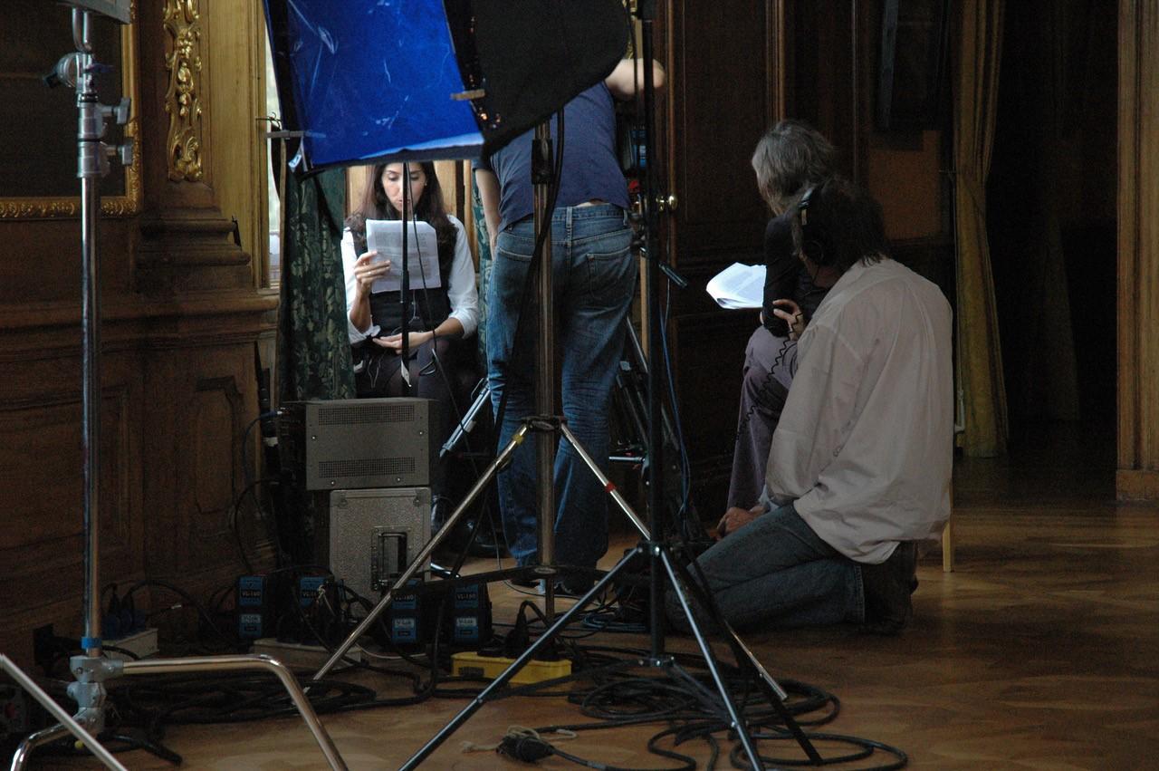 In einem Zimmer mit barocker Ausstattung sitzt eine Frau am Fenster und liest ein Manuskritp. Dabei wird sie gefilmt. Das sie umgebende Filmteam besteht aus drei Personen.