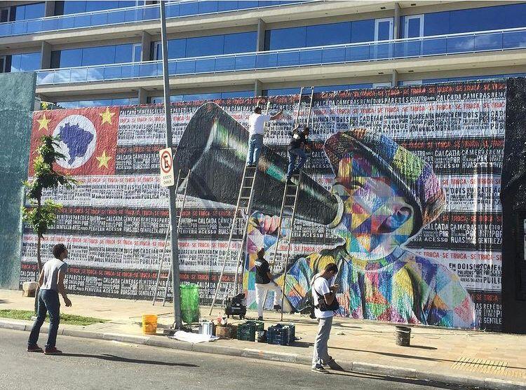 Menschen arbeiten an einem Street Art Werk. Es bedeckt eine Mauer vollständig und zeigt ein Kind mit Megaphone auf einer Kollage aus Plakaten.