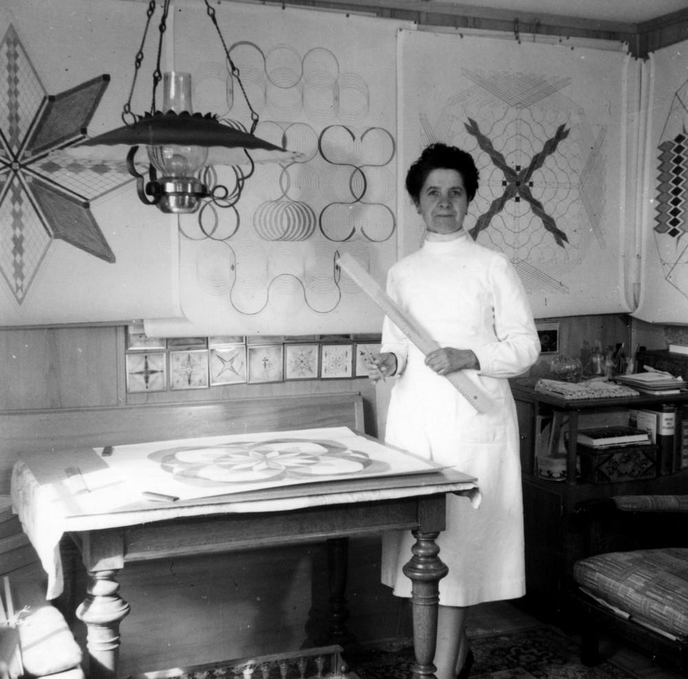 Emma Kunz steht vor einem Zeichentisch in Waldstatt. Sie trägt einen Kittel und hält ein langes Lineal und einen Stift in den Hand. An den Wänden hängen grafische Zeichnungen. Die Schwarzweißaufnahme entstand 1952 oder 53.