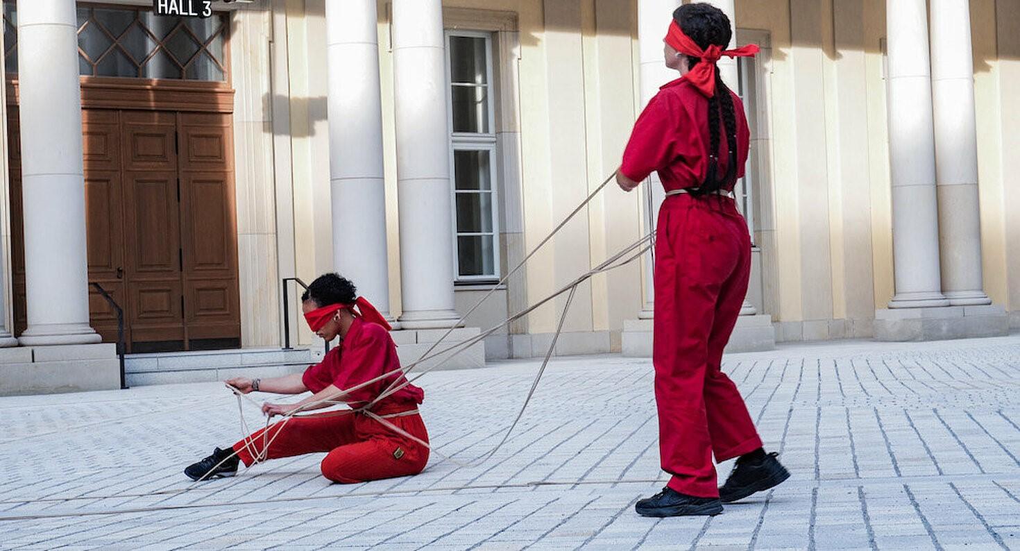 Zwei Menschen in roten Arbeitsanzügen und mit roten Augenbinden tanzen auf Abstand mit Seilen in einem säulenumstellten Innenhof.