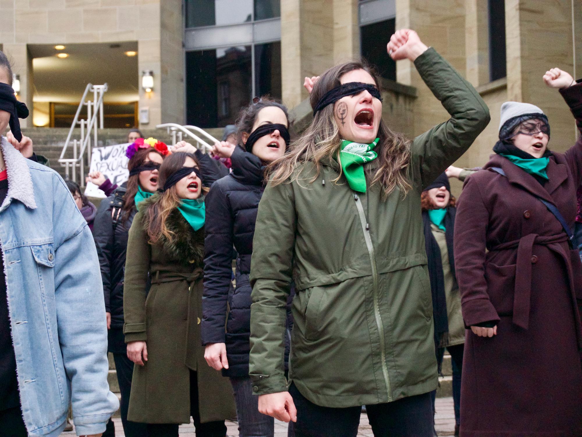 Protestierende Frauen auf der Straße, die teils schwarze Augenbinden tragen und eine Faust erheben. Die Aufnahme ist zeitgenössisch und wurde in Glasgow (UK) gemacht.